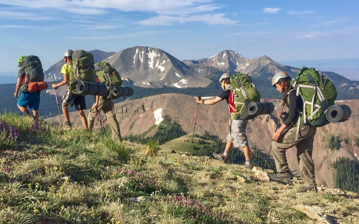 A group of students wearing backpacks hike along a trail. They appear to be at high elevation. There are mountains in the background. 